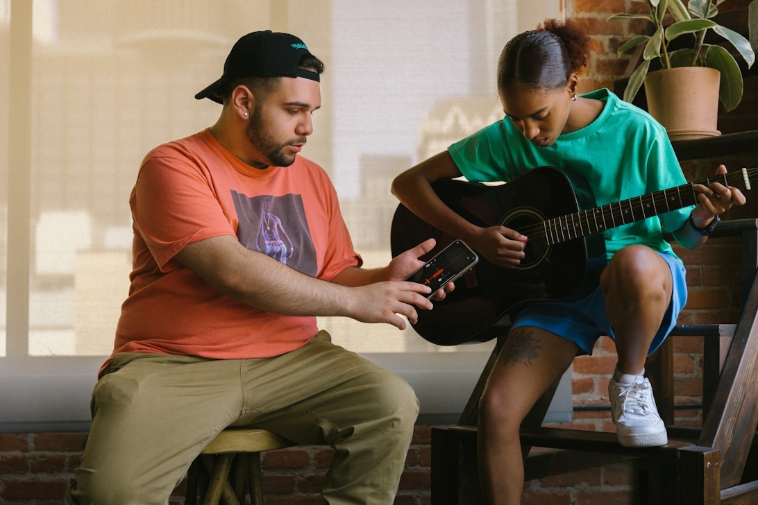Two musicians collaborating in a cozy indoor setting, with one person playing acoustic guitar while the other operates a mobile device, likely recording or sharing music ideas together.